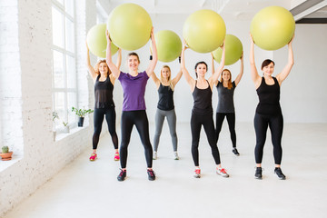 Fototapeta na wymiar Group of six young attractive sporty women holding green fitness balls above head while doing pilates or yoga exercise at white fitness studio interior. Team, friendship and helthy lifestyle concept.