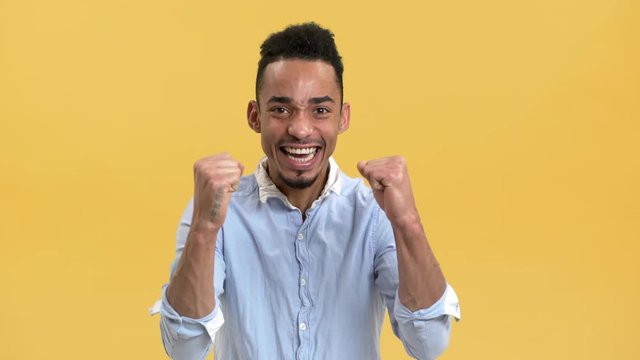 Portrait of delighted male student with arabic appearance rejoicing and clenching fists when passing exam, over yellow background. Concept of emotions