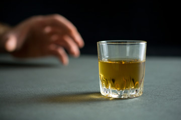 hand reaches for a glass of whiskey or cognac or alcohol drink, alcoholism and alcohol abuse concept, defocused, selective focus, close up, gray table, dark background