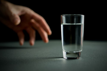 hand reaches for a glass of vodka or alcohol drink, alcoholism and alcohol abuse concept, defocused,.selective focus, close up, gray table, dark background