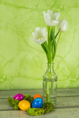 Drei weiße Tulpen in einer Vase aus Glas, davor einige bunte Ostereier mit Moos, grüner Hintergrund,