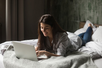 Smiling attractive woman lying on bed using laptop communicating online at home, happy girl typing...