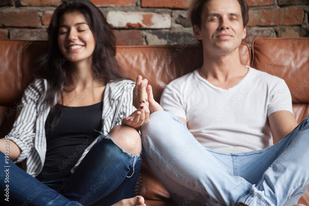 Wall mural young hipster couple practicing yoga pose together at home on sofa, smiling man and woman meditating