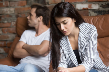 Sad pensive young girl thinking of relationships problems sitting on sofa with offended boyfriend,...