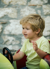 Emotional driver baby boy. Bright emotions while driving. Boy raised hand and shouted at wheel of toy car. Expressive emotions on child face. Screams and fun playing. Road and travel by car. Racing