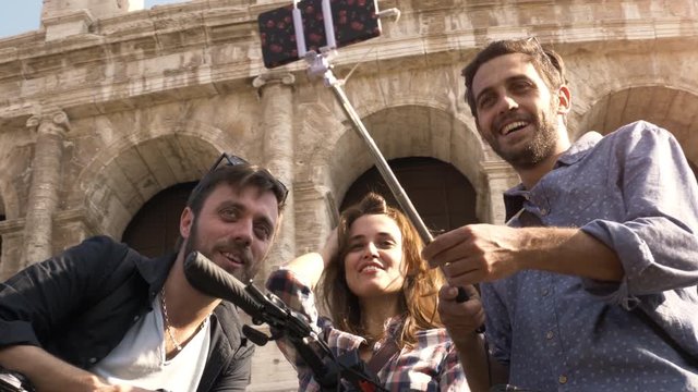 Three happy young friends tourists with bikes and backpacks at Colosseum in Rome taking selfies pictures with smartphone and stick having fun on sunny day slow motion steadycam