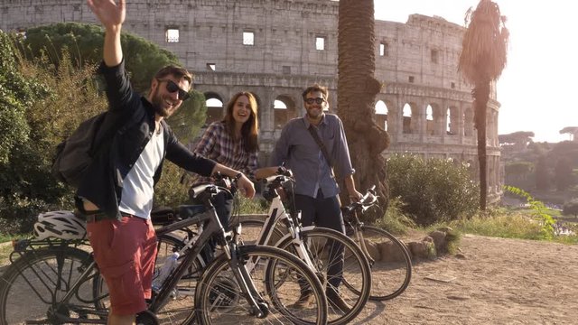 Three happy young friends tourists with bikes and backpacks at Colosseum in Rome waving hands to camera having fun at sunset on hill with trees slow motion