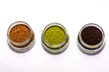 Cocoa powder, matcha tea powder, and ground coffee in little jars white background overhead photo


