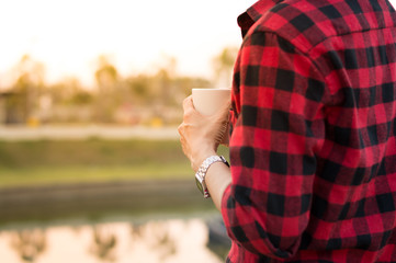 Man hold coffee cup in hand stand alone near the lake with morning light selective focus, copy space.