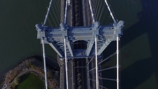 Overhead aerial view of the steel crown of a Manhattan Bridge tower where the main cables meet above the deck. The green river shore lies far below in a bold imposing industrial scene of New York.