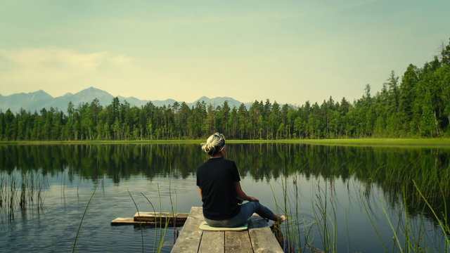 Lonely Dreaming Woman Sitting On A Wooden Bridge Pier Looking At The Mountains Reflected In The Mirror Water Of The Forest Lake. Adventure Nature Concept. Absolute Serenity