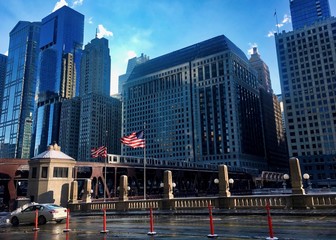 Elevated train passes over Chicago River as American flags fly in breeze.