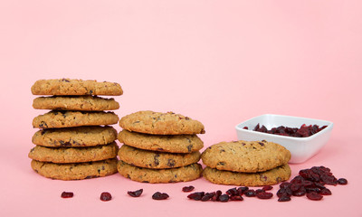 stacks of oatmeal cranberry cookies on a pink surface and background. Square bowl of dehydrated cranberries behind stacks with random cranberries on the table
