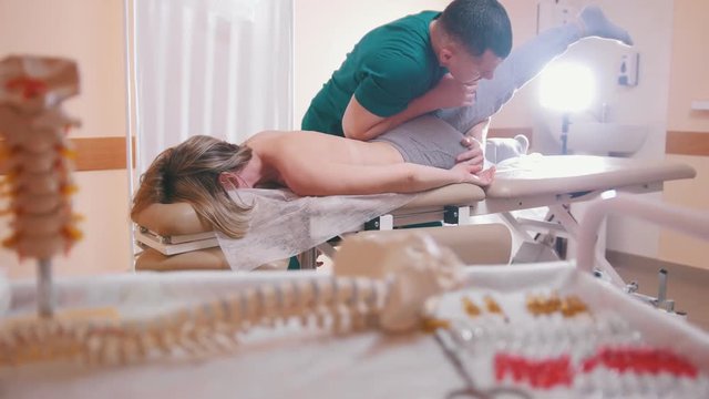 Chiropractor massaging a young woman lying on a massage table, stretching her joints with his elbow, spine model and drugs in the foreground