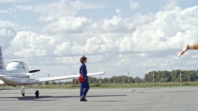Tracking shot of female aircraft mechanic in blue uniform walking on aerodrome at warm summer day