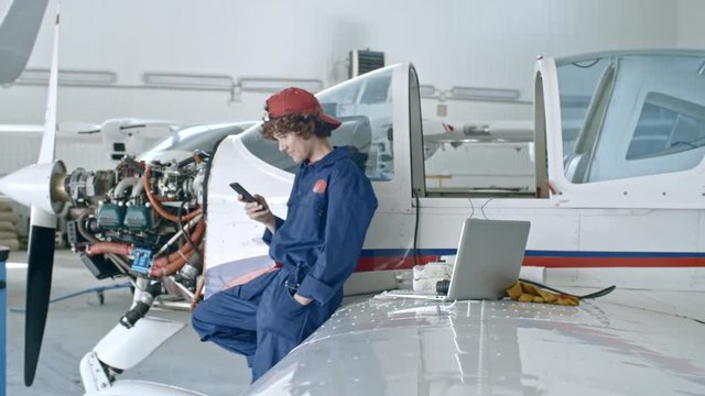 Young female aircraft mechanic standing by jet airplane in hangar, messaging on smartphone and smiling