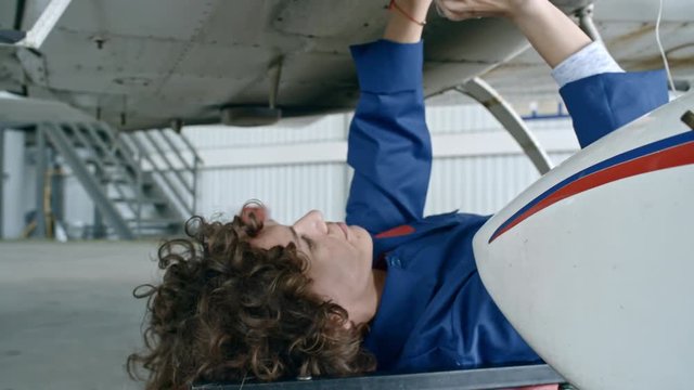 Zoom in of female aircraft mechanic lying on creeper underneath of airplane and fixing broken part in bottom