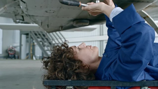 Female aircraft mechanic lying underneath of airplane and repairing bottom with screwdriver