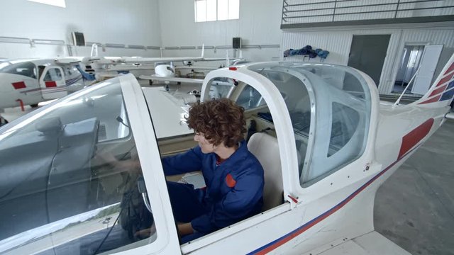 Female aviator in blue uniform getting into pilot cabin of light aircraft in hangar and checking cockpit before training flight