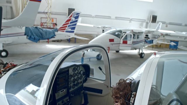 Tilt down of female pilot sitting in cabin of jet airplane in hangar, smiling and talking via headset with microphone