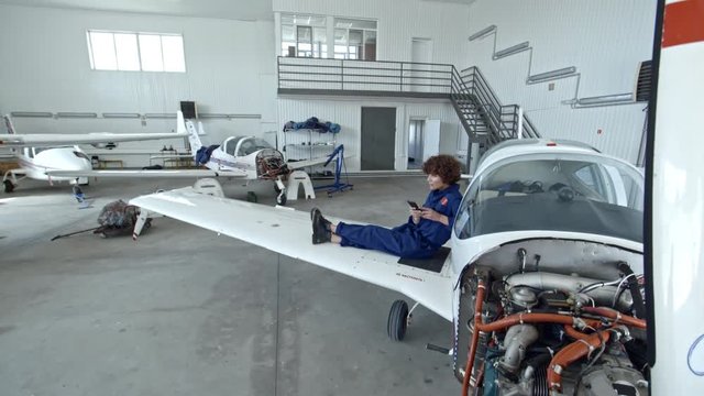 Tilt down of young female aircraft mechanic sitting on wing of jet airplane in hangar and texting on smartphone while having a break