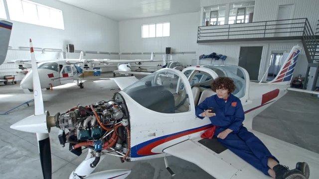 Tilt down of female aircraft mechanic resting on wing of jet airplane with exposed engine and texting on smartphone while having a break during workday in hangar