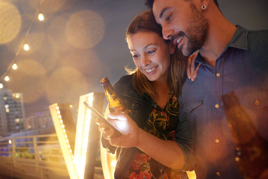 Couple having a drink at rooftop party and using smartphone