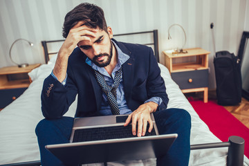 Working in hotel room. Confident young businessman in suit and tie working on laptop while sitting...