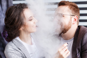 Couple vaping. Young man and woman blowing smoke at black and white striped studio background. Relationship and vape addiction concept with copy space. Close-up portrait