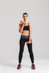 Full-length photo of sporty slim woman doing exercises with small dumbbells, isolated over gray background