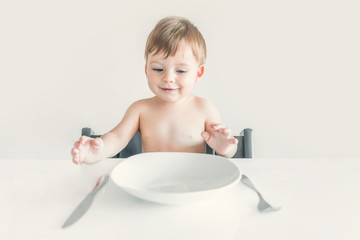 Hungry small blond boy sitting at the table with an empty white plate, fork and knife