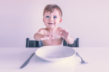 Smiling small blue-eyed blond boy sitting at the table with an empty white plate, fork and knife