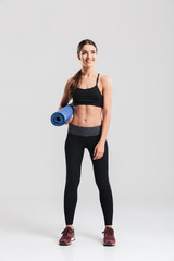 Full-length photo of healthy young woman in sportswear holding yoga mat while practising in studio, isolated over gray background