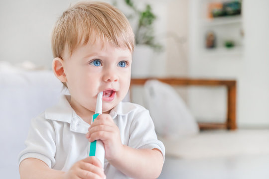 Small blue-eyed blond boy in a white polo shirt brushing his teeth with a toothbrush in the room
