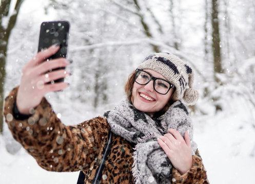 Attractive girl wears glasses making selfie by smartphone in winter snowy park