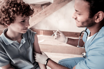 Brave patient. Side view on relaxed teenager and a physician having pleasant chat and smiling before vaccination indoors.