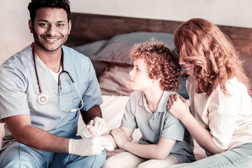 Always ready to help. Cheerful male medical worker grinning broadly into the camera while putting a bandage on an arm of a young patient.