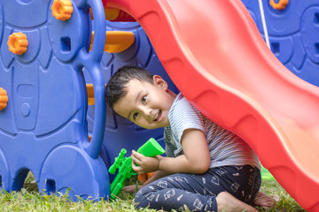 Portrait Thai boy hide under the slider with gun in the playground in front of the house. He is...