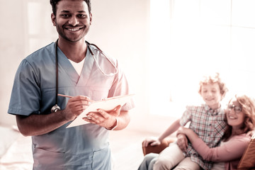 Positive attitude to work. Cheerful doctor looking into the camera with a broad smile on his face while holding a clipboard and making some notes.