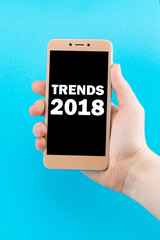 Trends 2018 inscription on phone screen. Golden phone in hand on a blue background