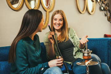 Two happy women are sitting in shisha bar and smoking nargile