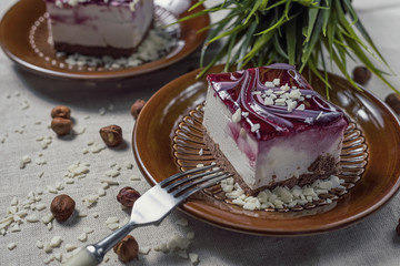 A piece of vanilla cake with chocolate base and cherry glaze decorated with some pieces of white chocolate served in a brown clay dish with silver fork placed on a beige tablecloth. Blurred background