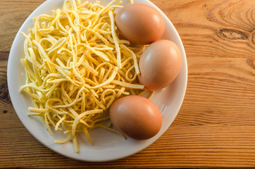 Three fresh chicken eggs, a pile of uncooked spaghetti noodles and a white plate on a wooden surface plateau, top view