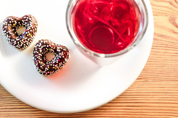 Two heart-shaped chocolate cookies with colorful candy sprinkles on top, glass of natural sour cherry red juice and a white plate on a wooden tabletop, top view
