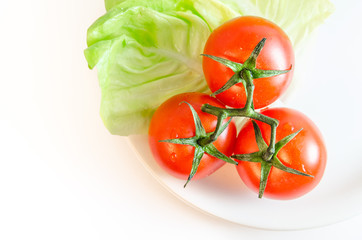Three cherry tomatoes with stems, green letuce leaves and a white plate, isolated on white tabletop background surface, top view