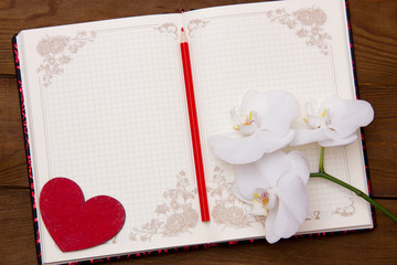 open blank notepad for notes with red pencil and red heart beside white orchid flowers on a wooden table