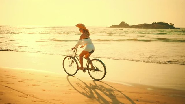 Woman in white and hat mooving on her bicycle along beach sand at summer time under burning sunset sky, lifestyle carefree.
