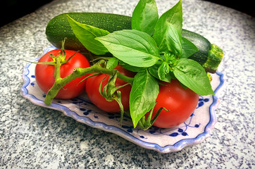 Ceramic plate with ripe red tomatoes, zucchini and green leaves of basil, on a marble blurred table