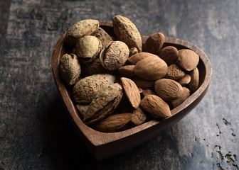 Raw almonds with and without its shell in a wooden heart-shaped bowl. Dark rustic wood background, selective focus
