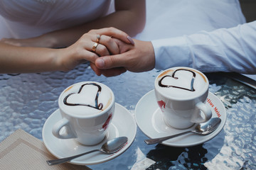 Wedding couple is sitting and holding arms. Nice cappuccino with foam and hearts on the table. Glass table and engagement golden ring.